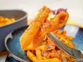 Widok z bliska na widelcu, na którym znajduje się makaron penne z sosem pomidorowym i kawałkami chorizo.