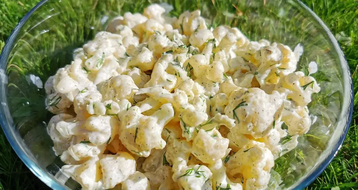 Pyszna sałatka z kalafiora w kremowym sosie z koperkiem, podana w szklanej misie na tle zielonej trawy.