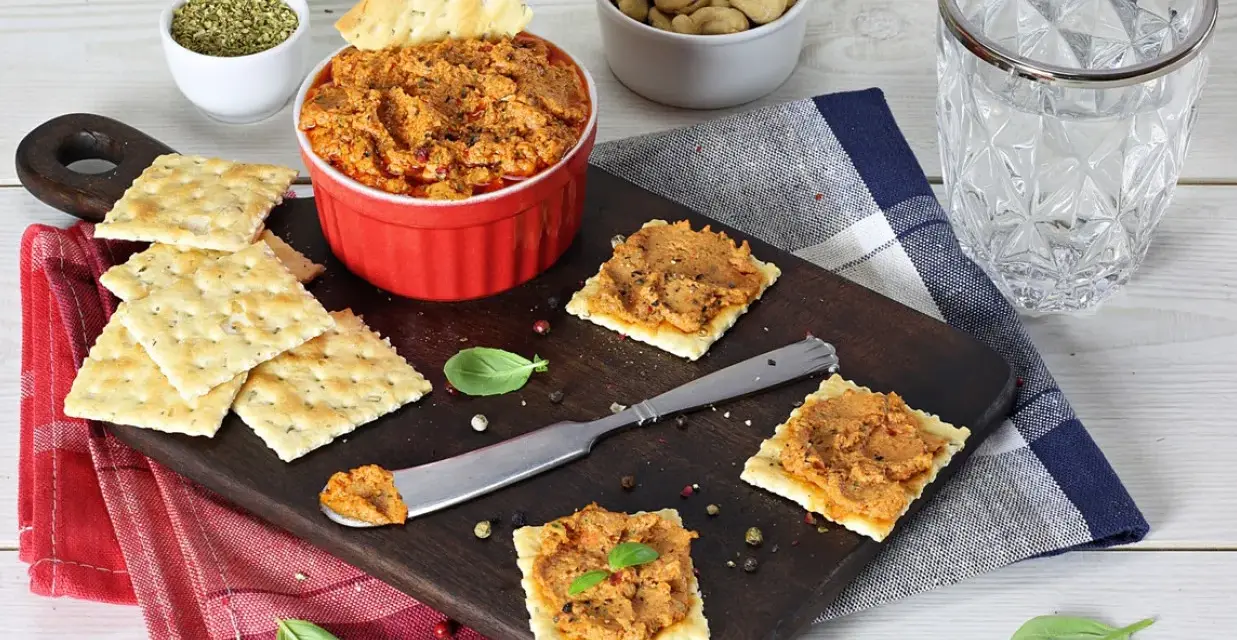 Pyszna pasta z suszonych pomidorów na krakersach, gotowa do degustacji. Obok miseczka z orzechami i szklanka wody.