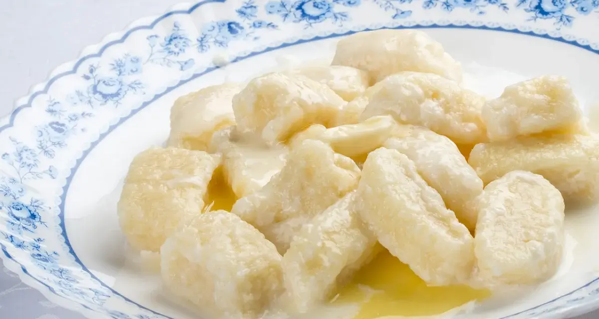 Pyszne kopytka z serem, polane śmietaną i masłem, na talerzu z niebieskim wzorem.