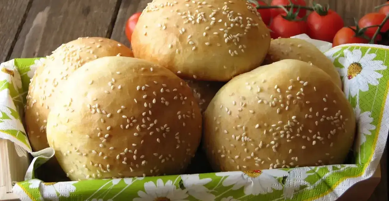Świeżo upieczone bułki do burgerów, posypane sezamem, czekają w koszyku. Obok gałązka pomidorków koktajlowych.