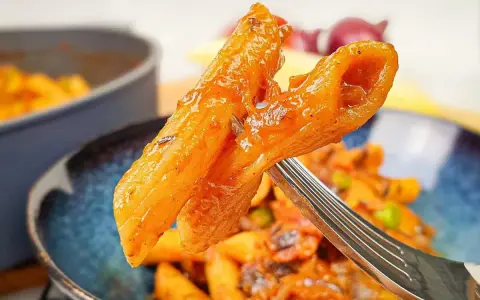 Widok z bliska na widelcu, na którym znajduje się makaron penne z sosem pomidorowym i kawałkami chorizo.