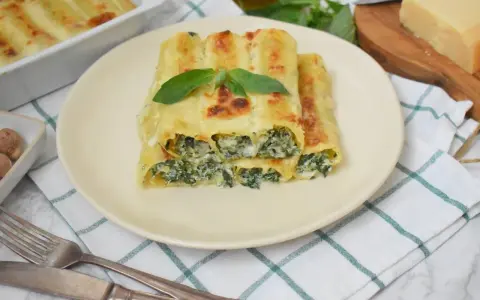Pyszne cannelloni ze szpinakiem, zapieczone pod beszamelem i serem, podane na talerzu z listkiem bazylii.