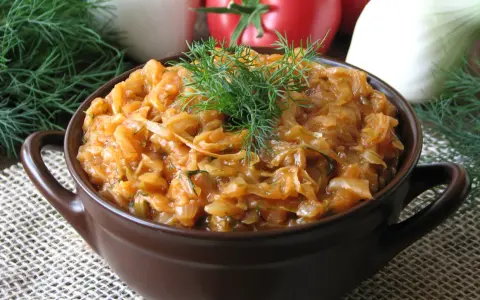 Młoda kapusta z pomidorami, duszona z koperkiem, w brązowej miseczce. W tle pomidory i cebula.