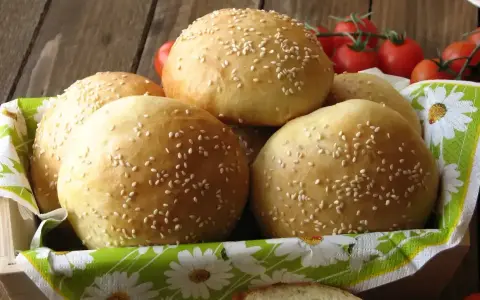 Świeżo upieczone bułki do burgerów, posypane sezamem, czekają w koszyku. Obok gałązka pomidorków koktajlowych.