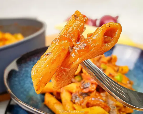 Widok z bliska na widelcu, na którym znajduje się makaron penne z sosem pomidorowym i kawałkami chorizo.