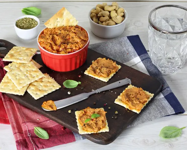 Pyszna pasta z suszonych pomidorów na krakersach, gotowa do degustacji. Obok miseczka z orzechami i szklanka wody.