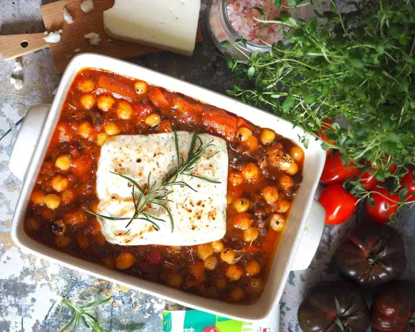 Pyszny gulasz z ciecierzycy w sosie pomidorowym, zapieczony z serem feta i gałązką rozmarynu. Obok świeże pomidory i zioła.