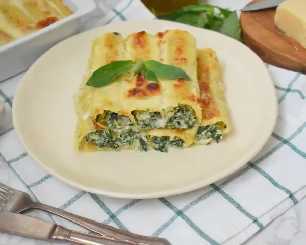 Pyszne cannelloni ze szpinakiem, zapieczone pod beszamelem i serem, podane na talerzu z listkiem bazylii.