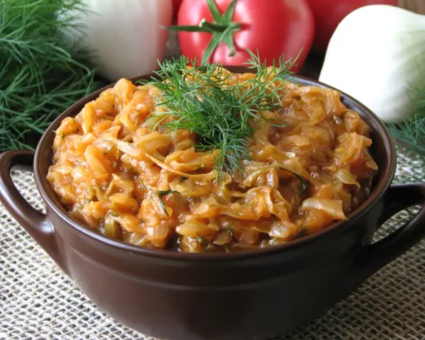 Młoda kapusta z pomidorami, duszona z koperkiem, w brązowej miseczce. W tle pomidory i cebula.