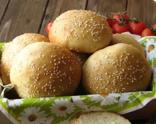 Świeżo upieczone bułki do burgerów, posypane sezamem, czekają w koszyku. Obok gałązka pomidorków koktajlowych.