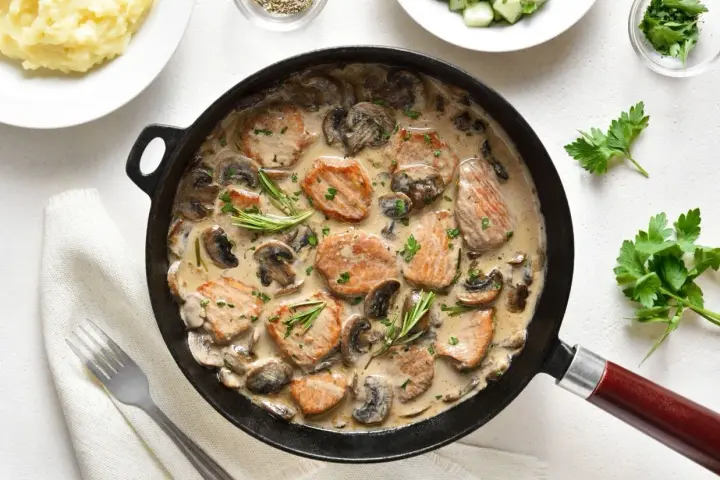 Pyszne polędwiczki w sosie pieczarkowym, podane w patelni, z gałązkami rozmarynu i posypane natką pietruszki. Obok puree ziemniaczane.