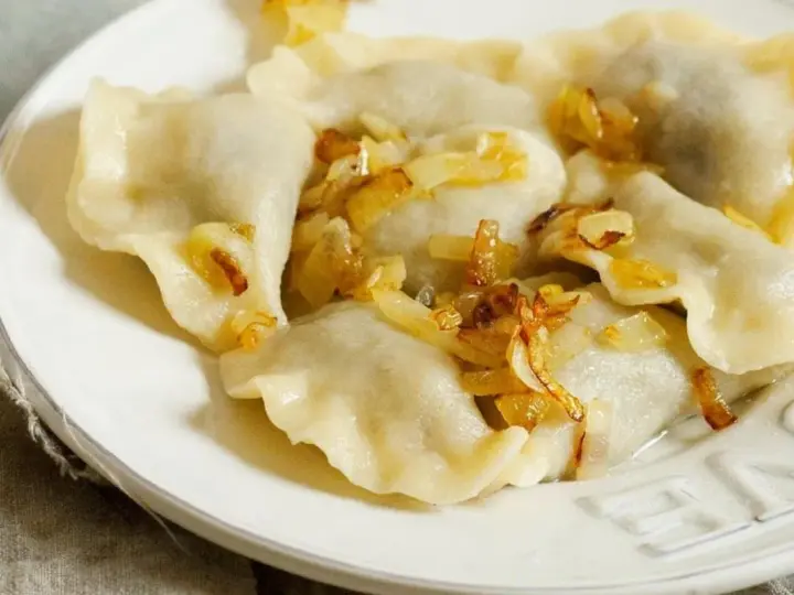 Pyszne pierogi z mięsem, polane skwarkami z cebulki, na białym talerzu.