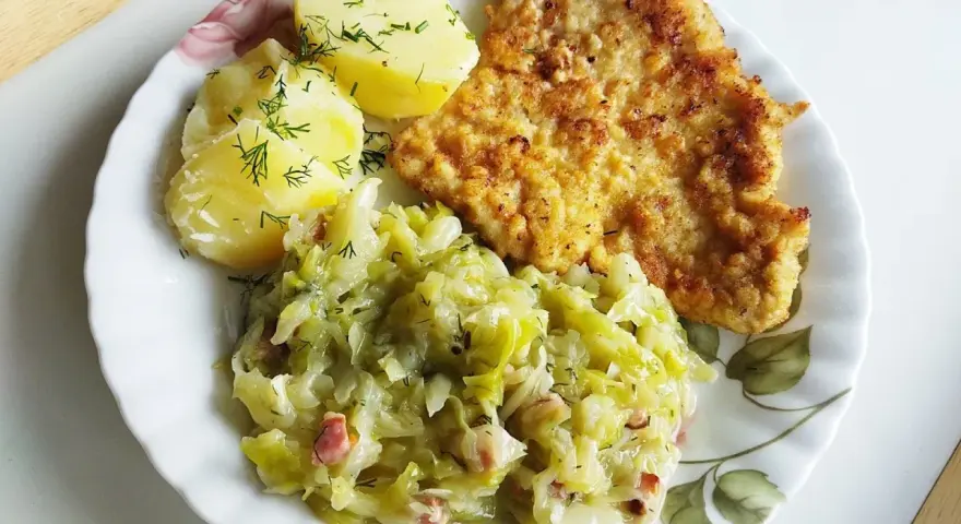 Pyszna kapusta zasmażana z boczkiem, podana z ziemniakami i kotletem.