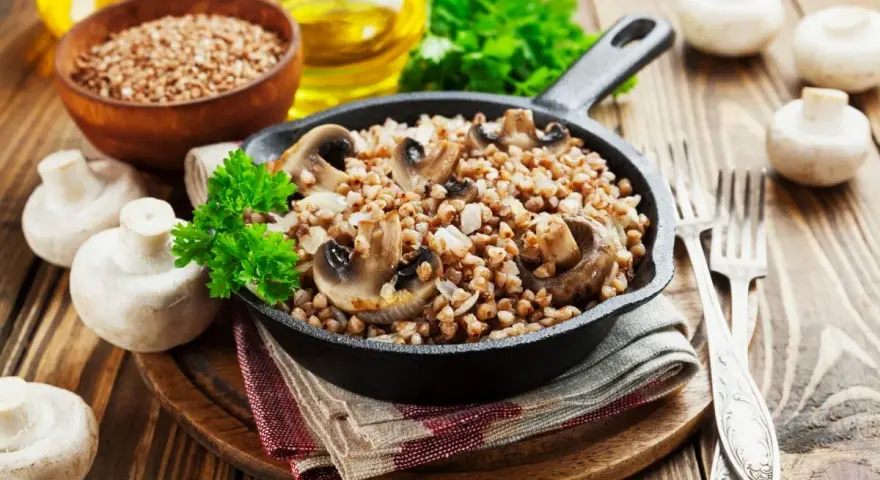 Pyszne kaszotto z pieczarkami w żeliwnej patelni, udekorowane natką pietruszki. Obok świeże pieczarki i miseczka kaszy gryczanej.