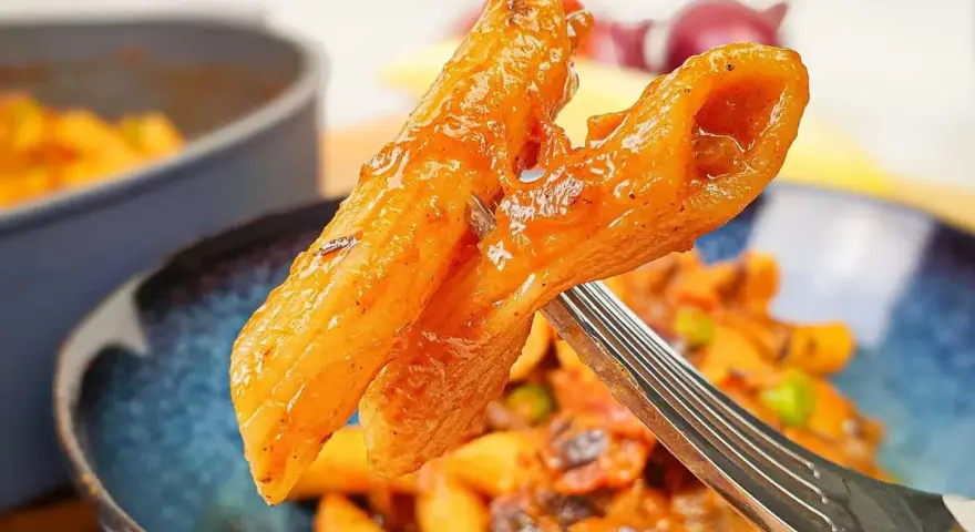 Widok z bliska na widelcu, na którym znajduje się makaron penne z sosem pomidorowym i kawałkami chorizo.