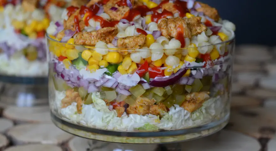 Warstwowa sałatka gyros z makaronem, kurczakiem, kukurydzą i warzywami w szklanej misie.