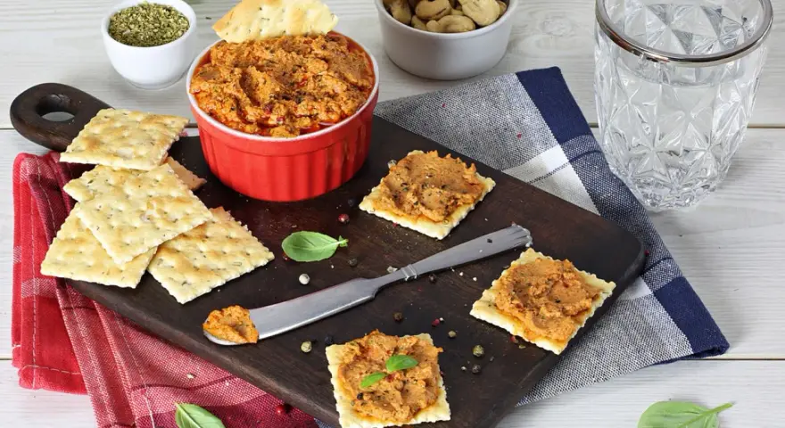 Pyszna pasta z suszonych pomidorów na krakersach, gotowa do degustacji. Obok miseczka z orzechami i szklanka wody.
