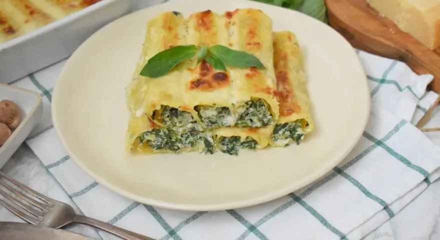 Pyszne cannelloni ze szpinakiem, zapieczone pod beszamelem i serem, podane na talerzu z listkiem bazylii.