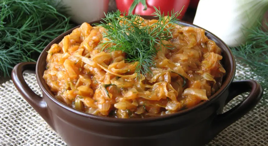 Młoda kapusta z pomidorami, duszona z koperkiem, w brązowej miseczce. W tle pomidory i cebula.