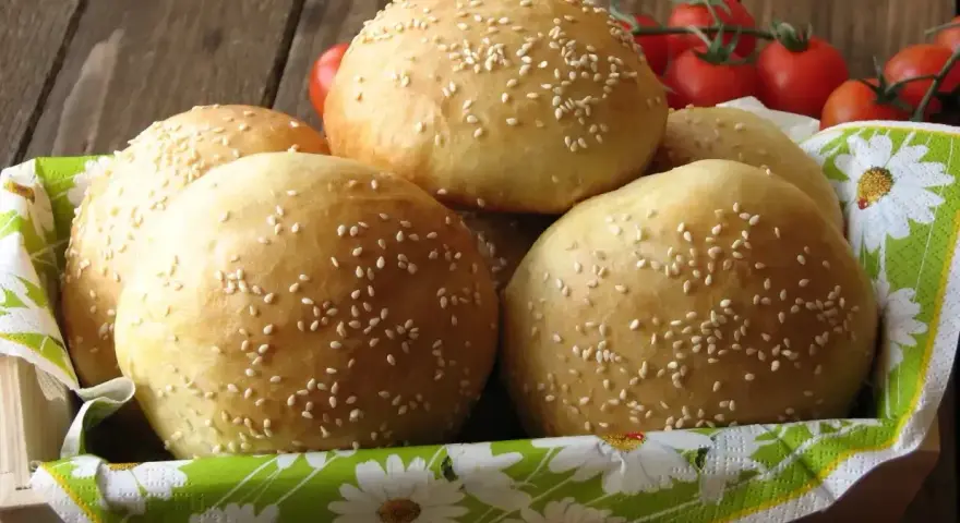 Świeżo upieczone bułki do burgerów, posypane sezamem, czekają w koszyku. Obok gałązka pomidorków koktajlowych.