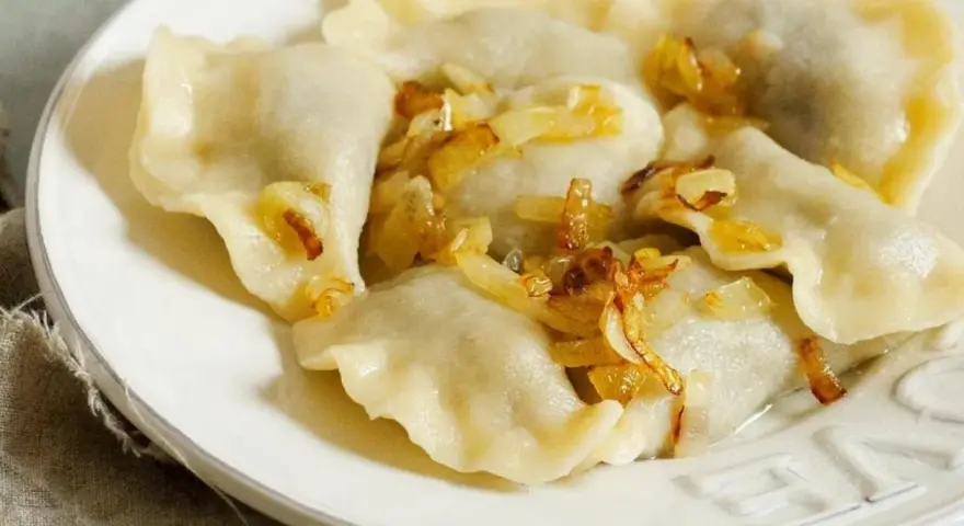 Pyszne pierogi z mięsem, polane skwarkami z cebulki, na białym talerzu.