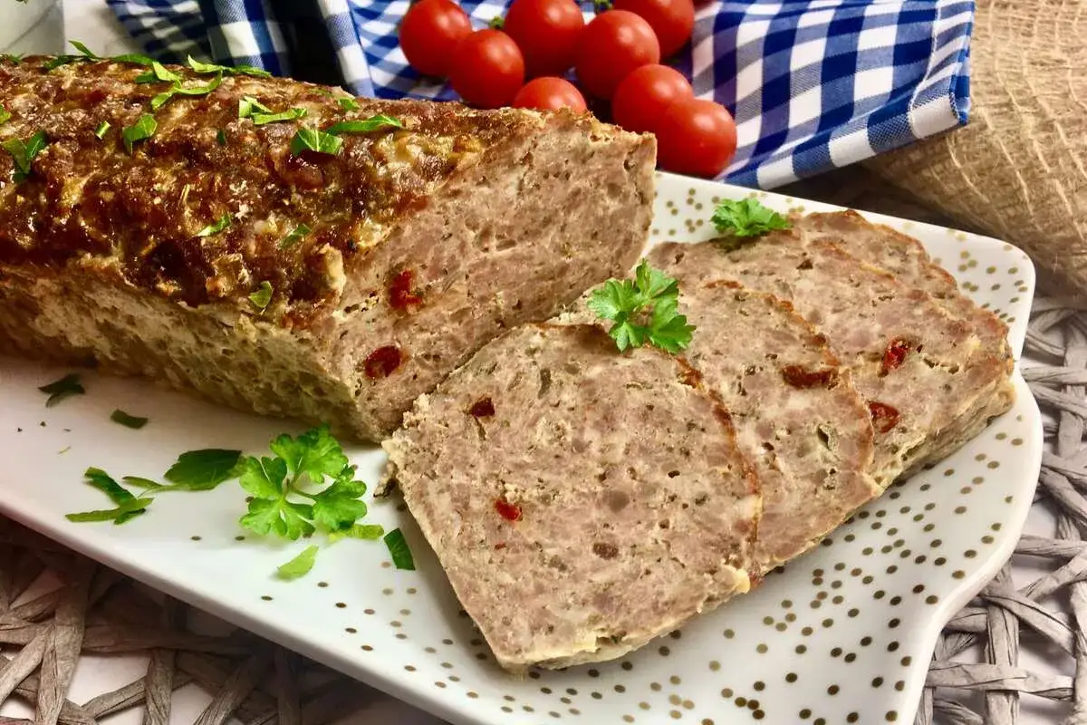 Pyszna pieczeń z mięsa mielonego, pokrojona w plastry, ozdobiona natką pietruszki. Obok wiśniowe pomidorki i niebiesko-biała kratka.
