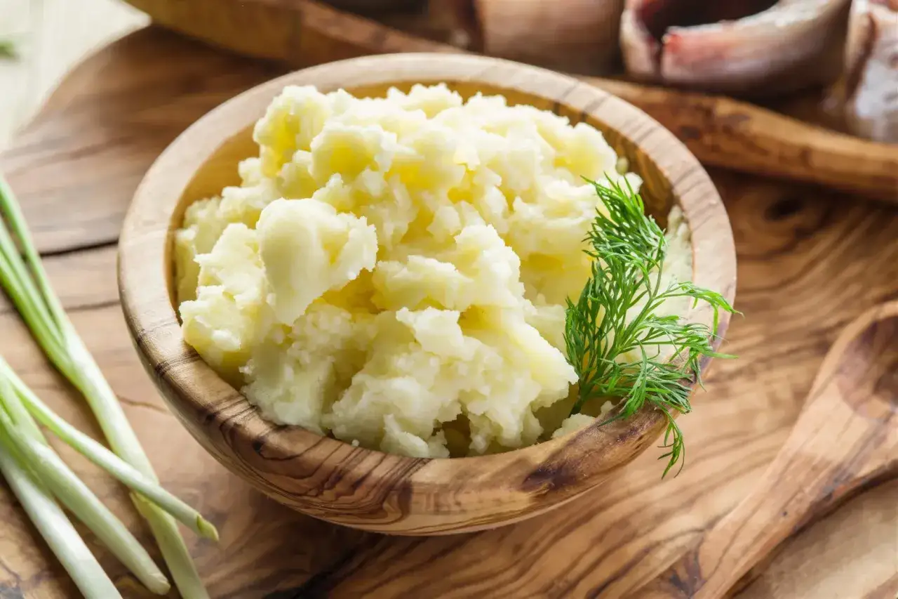 Puszyste puree ziemniaczane w drewnianej misce, obok szczypiorek i koperek. Idealne, by dowiedzieć się, jak zrobić puree.