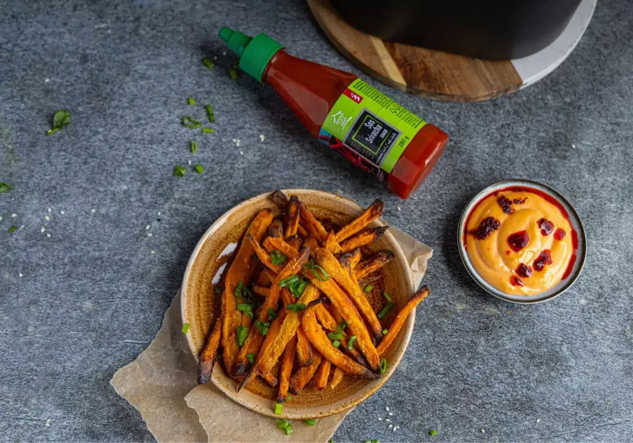 Chrupiące frytki z batatów z airfryera, posypane szczypiorkiem, obok sos sriracha i dip.