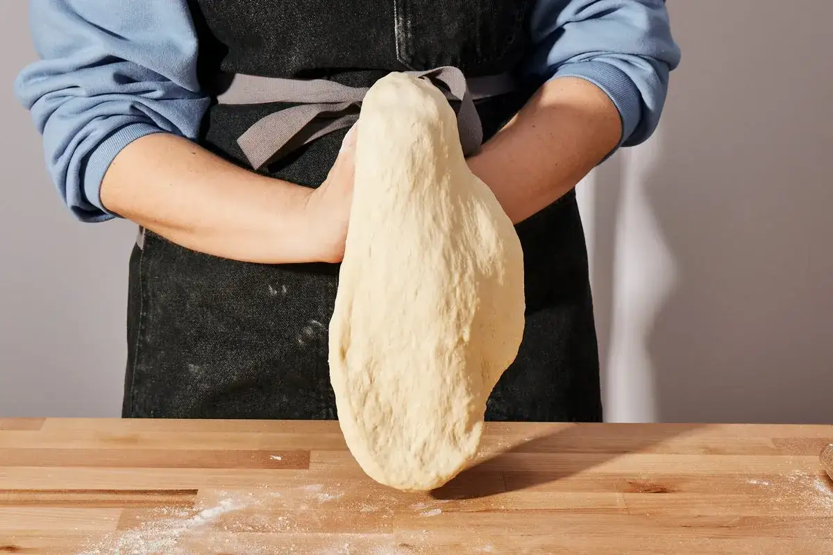 Osoba w fartuchu rozciąga elastyczne ciasto na pizzę nad drewnianym blatem.
